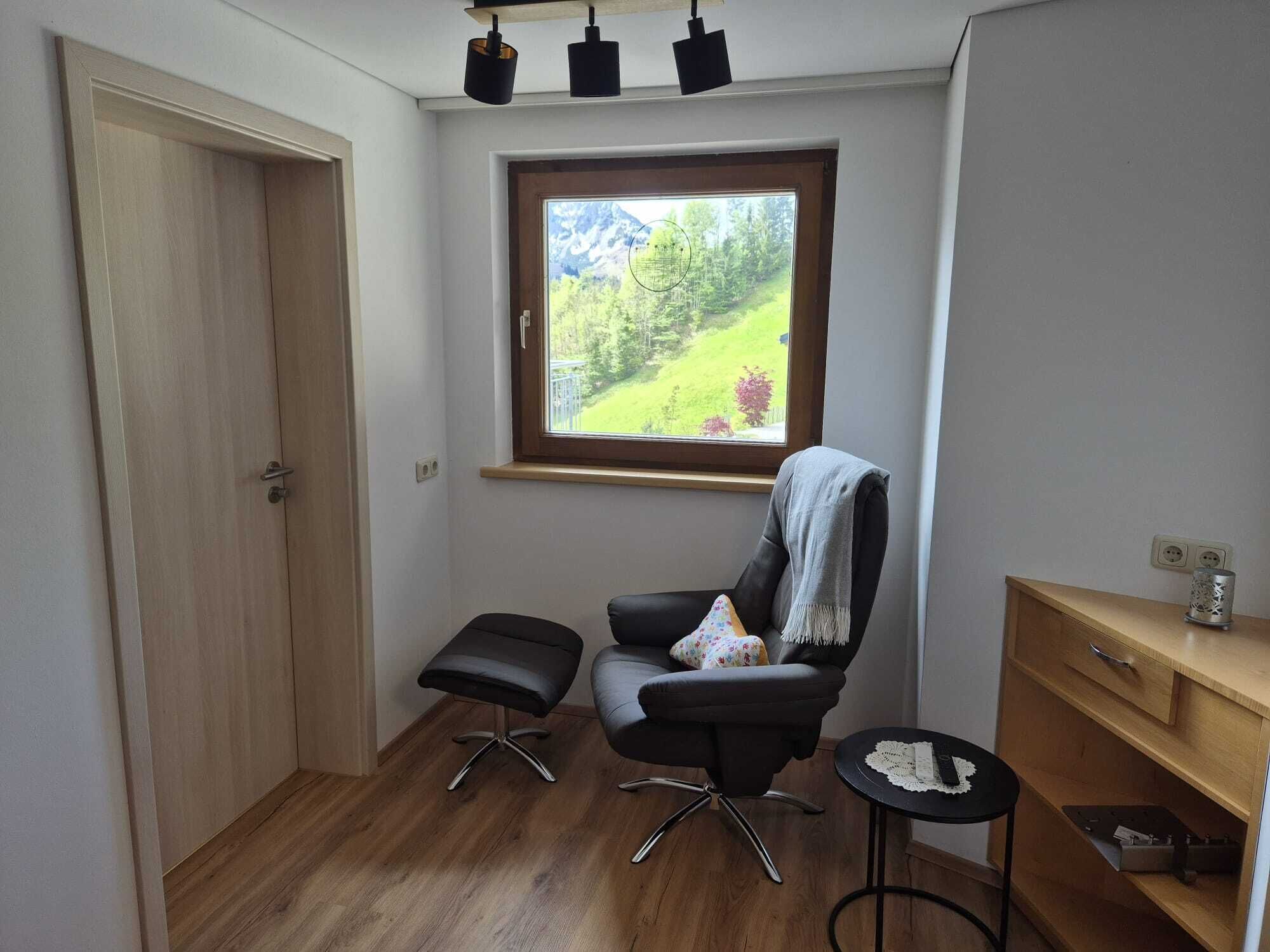 Une petite pièce bien éclairée, avec un plancher en bois, une chaise noire dotée d’un coussin, une petite table ronde et une fenêtre donnant sur un paysage verdoyant.