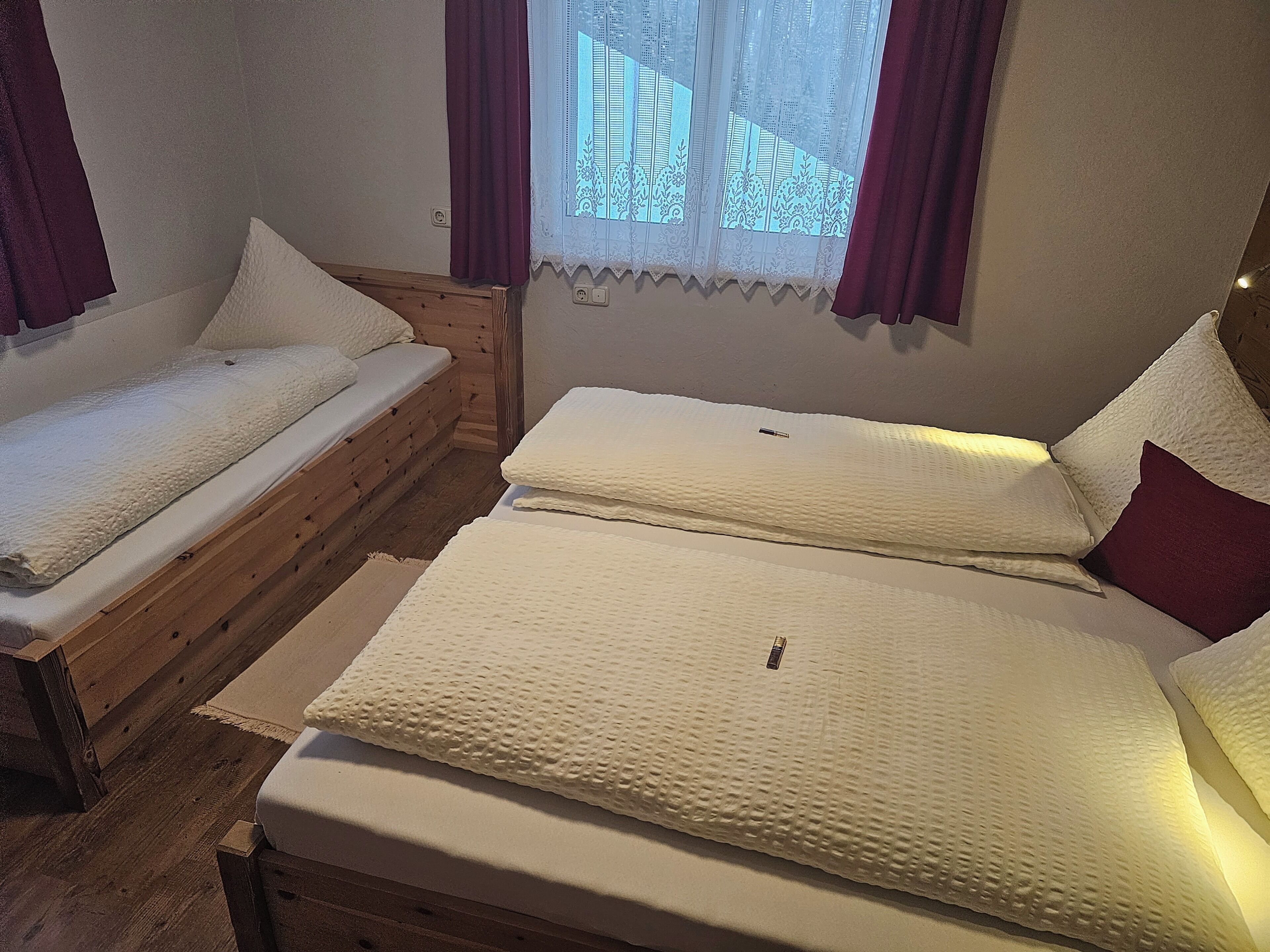 Quarto com duas camas, cabeceira de madeira, janela com cortinas de renda e um interruptor na parede.