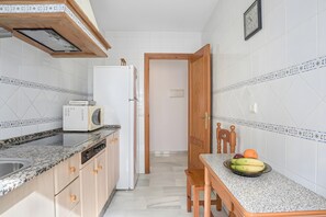 A compact kitchen with a microwave, toaster, and a wooden dining table with fruit. - Tomy Apartment – Families Only, Sea View, Balcony, Wi-Fi (Conil de la Frontera)