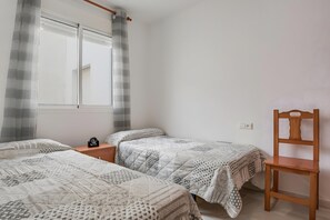 Ein Schlafzimmer mit zwei Einzelbetten, einem Holzstuhl, einem Nachttisch mit einer Uhr und einem Fenster mit Vorhängen.