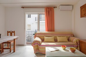 A small living room with a striped sofa, a dining table, and a window with curtains. - Tomy Apartment – Families Only, Sea View, Balcony, Wi-Fi (Conil de la Frontera)
