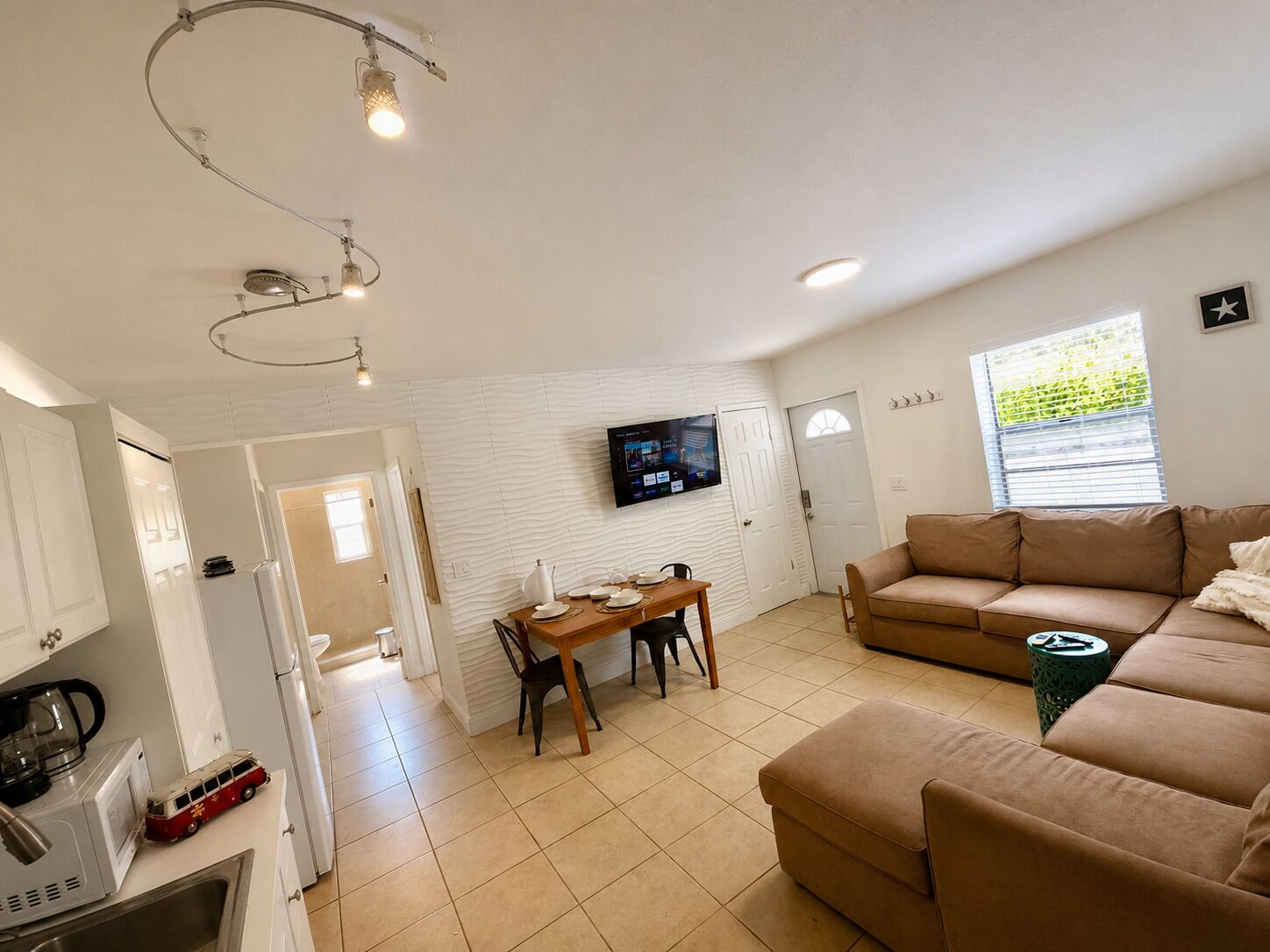 Ein modernes Wohnzimmer mit einem beigefarbenen Sofa, einem Essbereich mit Tisch und Stühlen, einer Küche mit Kühlschrank und Mikrowelle sowie einem an der Wand montierten Fernseher.