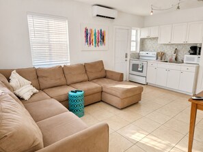 Un salon avec un canapé d’angle beige, un coin cuisine doté de meubles blancs et un pouf bleu à motifs.