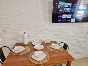 Ein Esszimmer mit Tisch, Geschirr, Besteck und Servietten. Im Hintergrund eine strukturierte Wand und ein Flachbildfernseher, der verschiedene Streaming-Dienste zeigt.