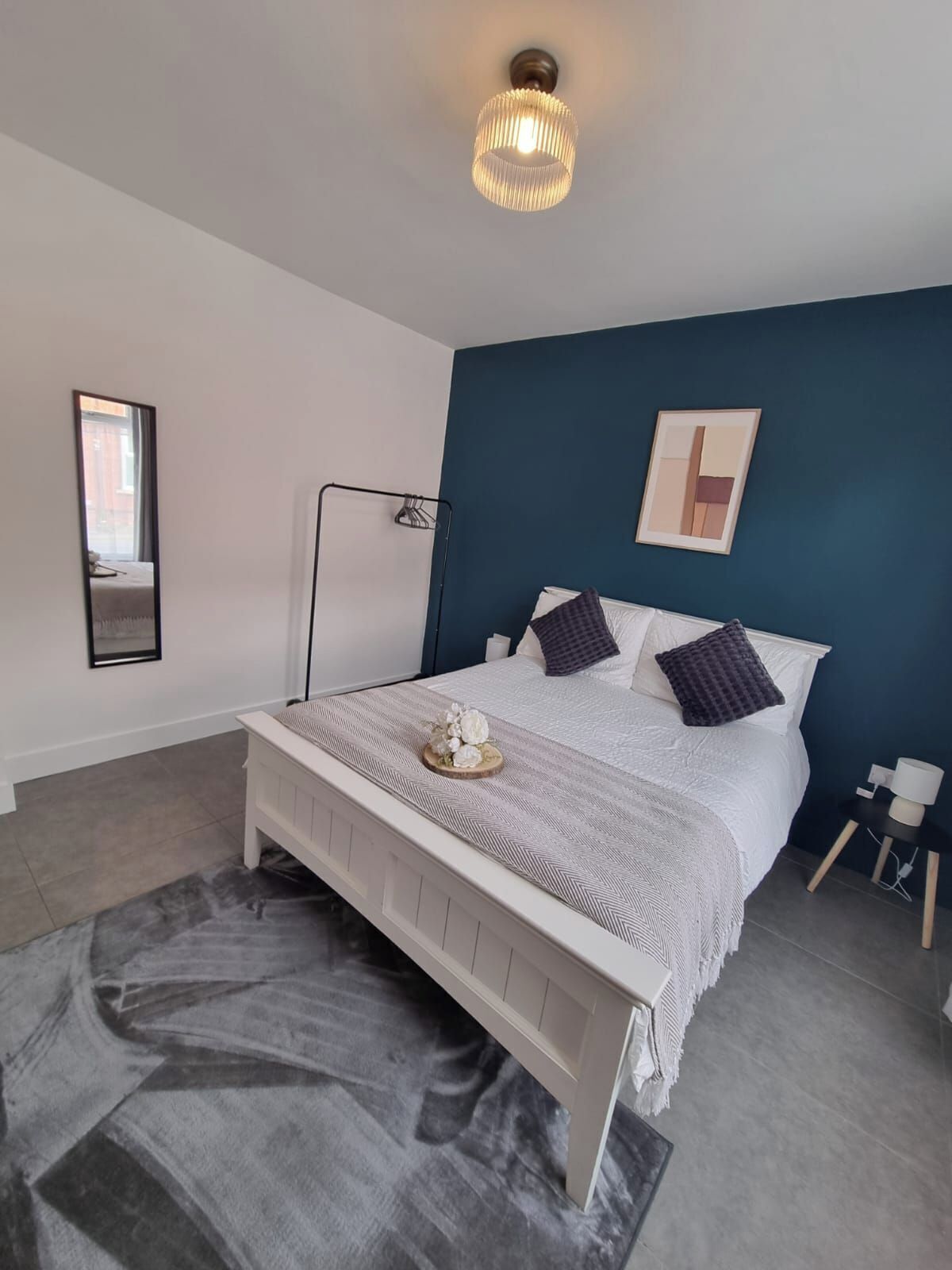 A bedroom with a bed, a bedside table, a lamp, a mirror, and a wall art piece.