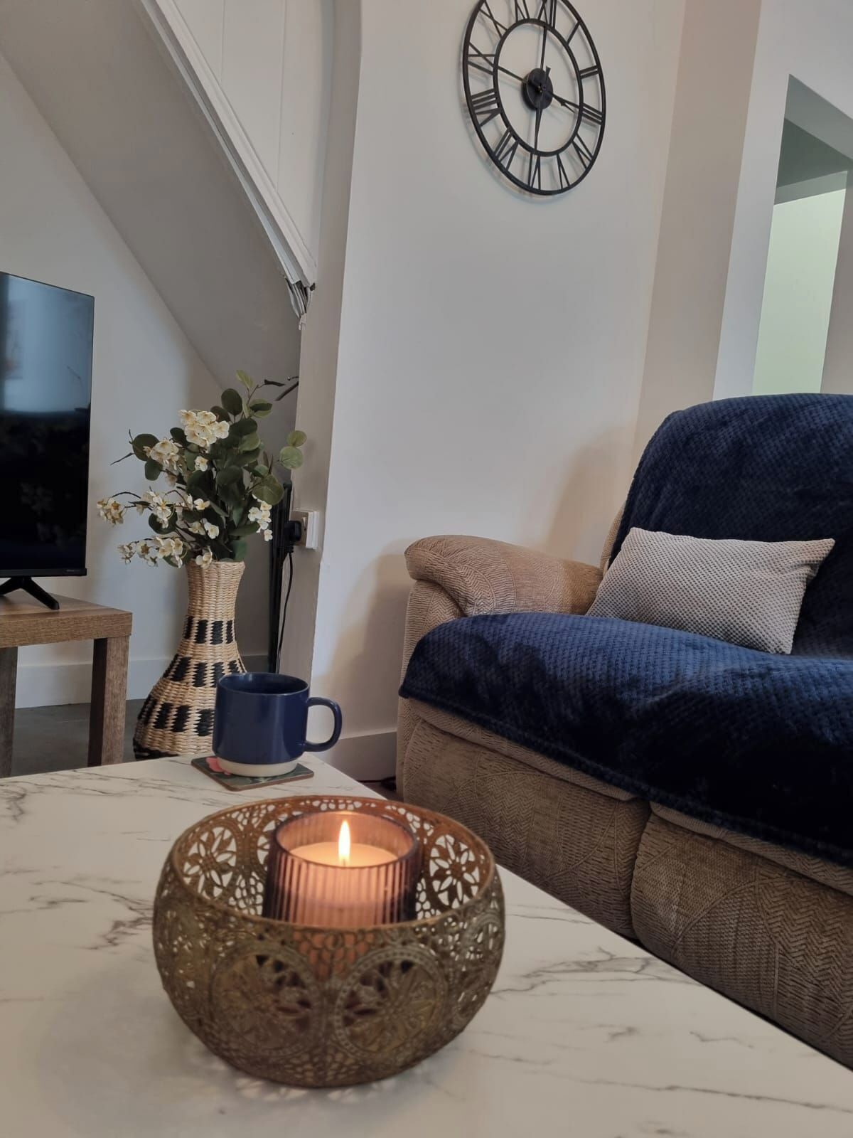 Un salon avec un canapé, une horloge murale, un téléviseur, un vase de fleurs et une bougie posée sur une table.