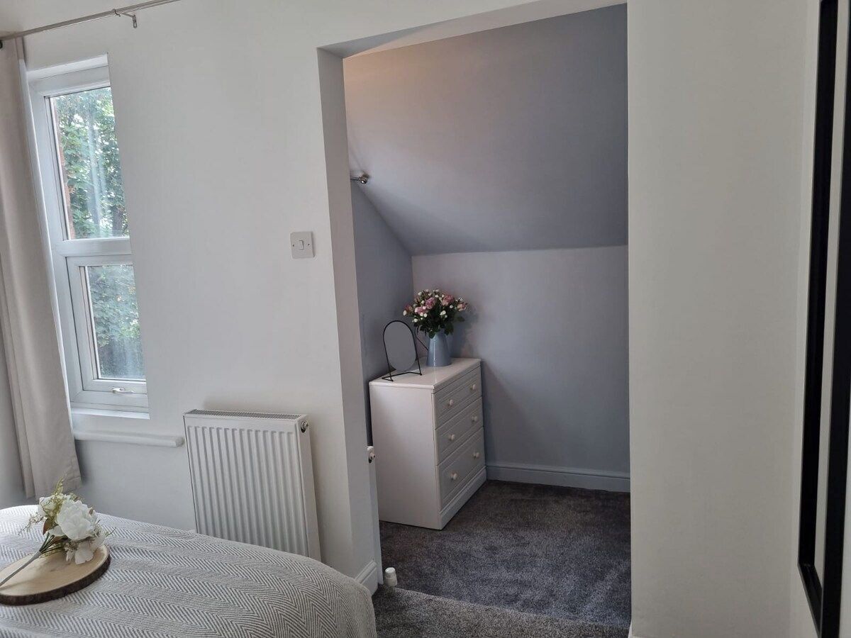 Et soveværelse med vindue, radiator, kommode og et lille bord med blomster.