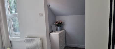 Une chambre avec une fenêtre, un radiateur, une commode et une petite table ornée de fleurs.