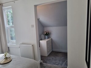 Une chambre avec une fenêtre, un radiateur, une commode et une petite table ornée de fleurs.