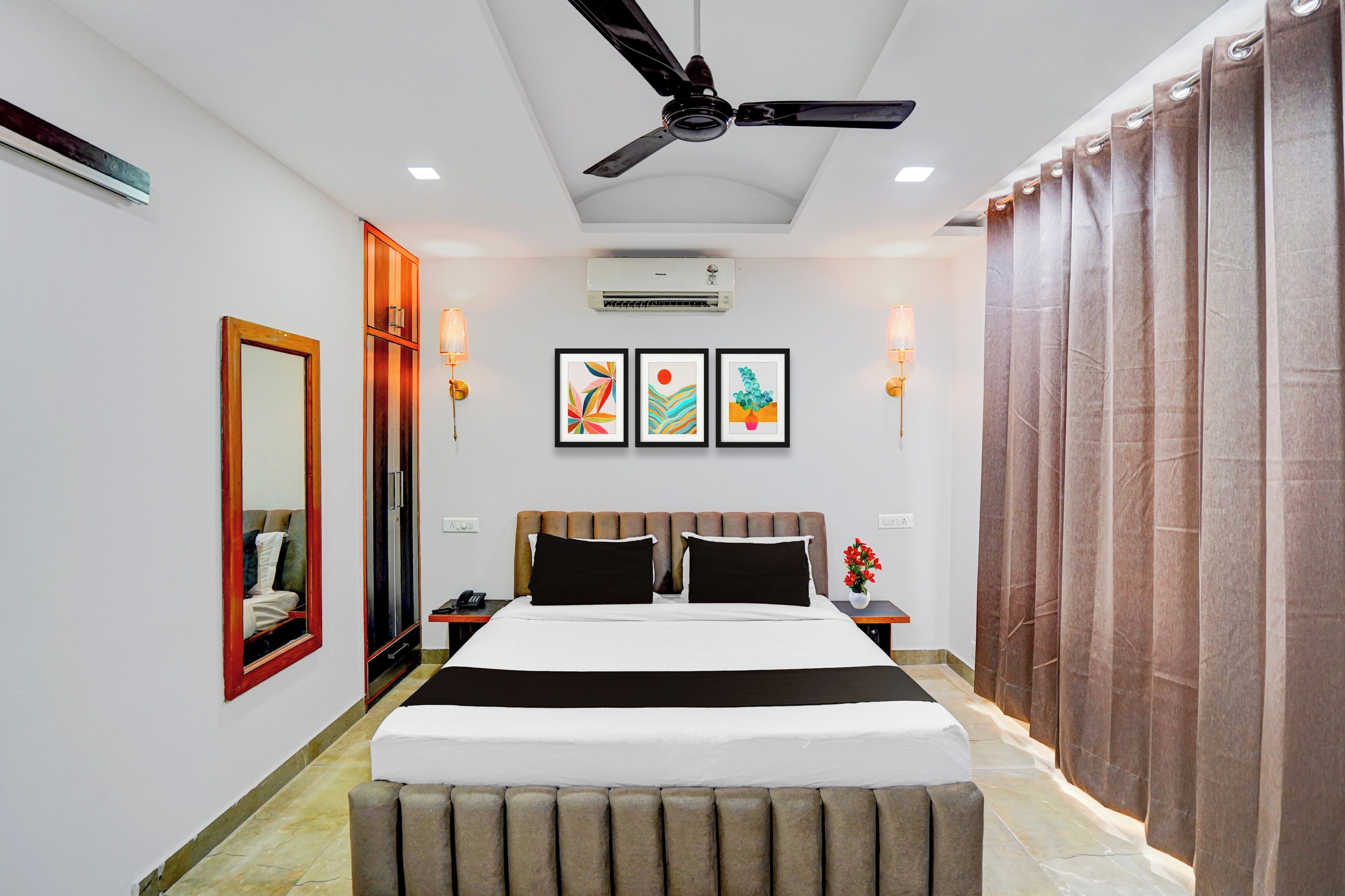 Una habitación de hotel con una cama, ventilador de techo, equipo de aire acondicionado, mesita de noche y una lámpara en la mesita auxiliar, junto a un ventanal con cortinas.