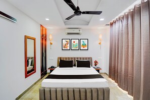 A hotel room with a bed, a ceiling fan, an air conditioning unit, a nightstand, a bedside table with a lamp, and a large window with curtains. - Super Townhouse by Cosmo Mall Zirakpur (Zirakpur)