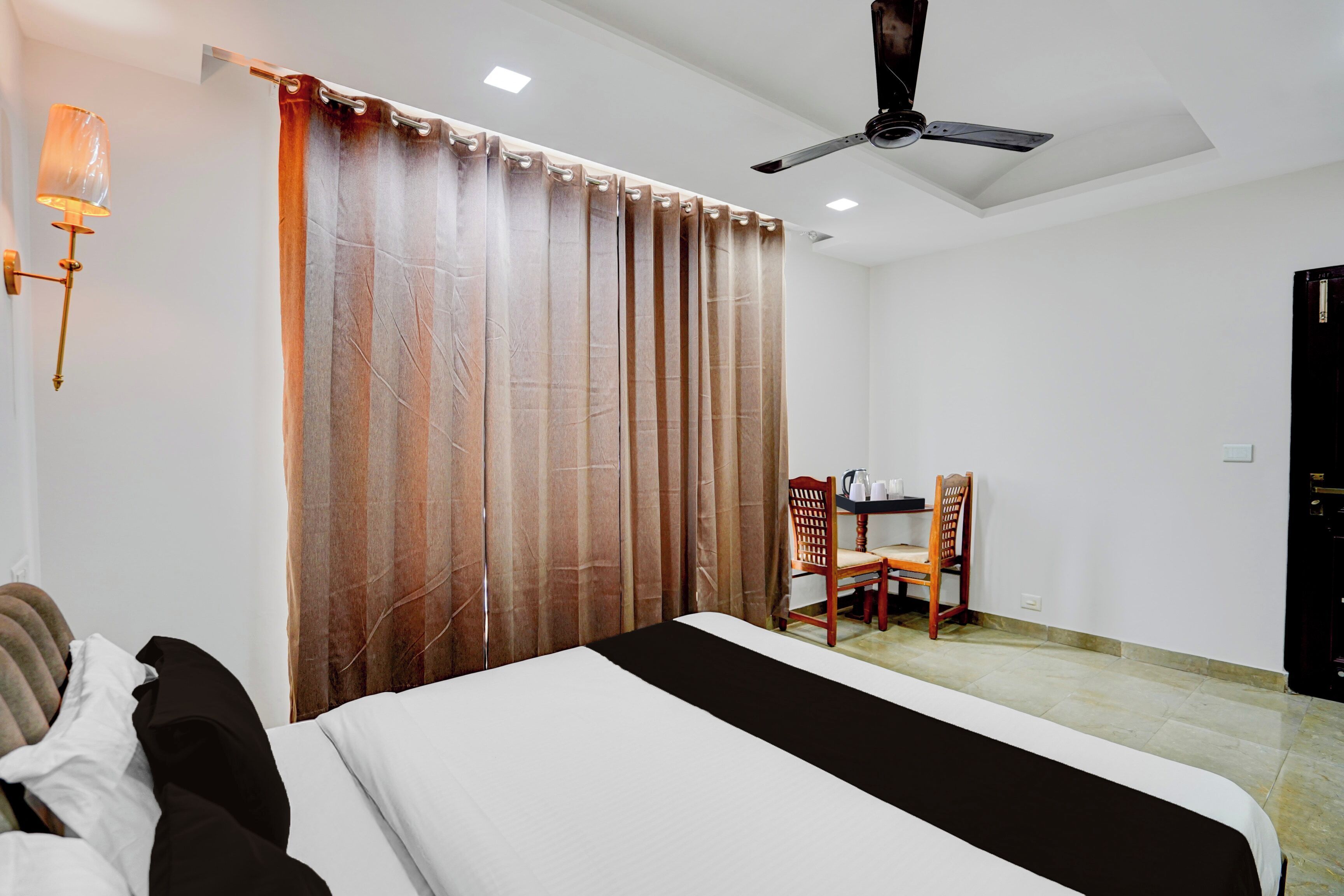 Habitación de hotel con cama, escritorio con sillas, ventilador de techo y lámpara de pared.