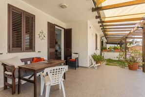 Een terras buiten met een houten tafel en stoelen, een pergola en potplanten.