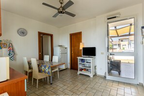 A room with a dining table, chairs, a ceiling fan, a television on a stand, a refrigerator, and a door leading outside. - Holiday Home 'Sul Porto – Steps from the Sea' with Sea View, Private Terrace & AC (Grosseto)