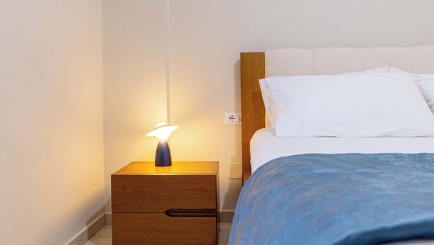 A hotel room with a wooden nightstand, a lamp, a bed with white bedding, and a blue blanket. - Villa Verde (Aliko)