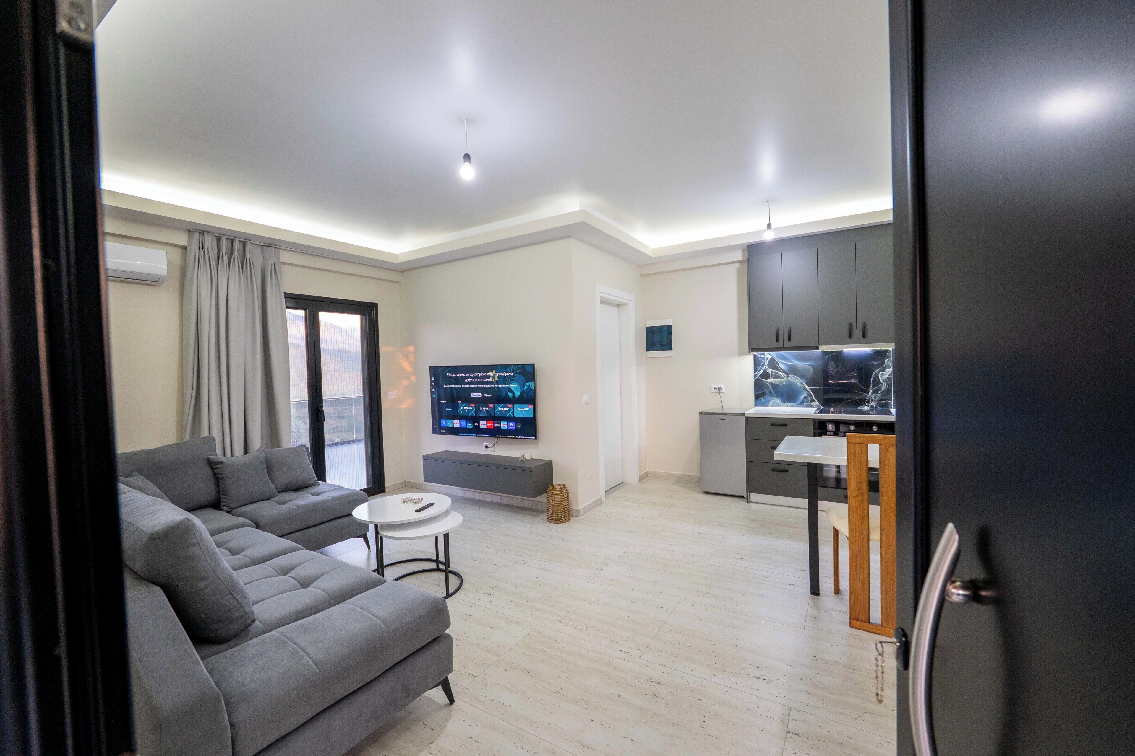 A modern living room with a grey sofa, a small round table, and a flat-screen TV mounted on the wall.