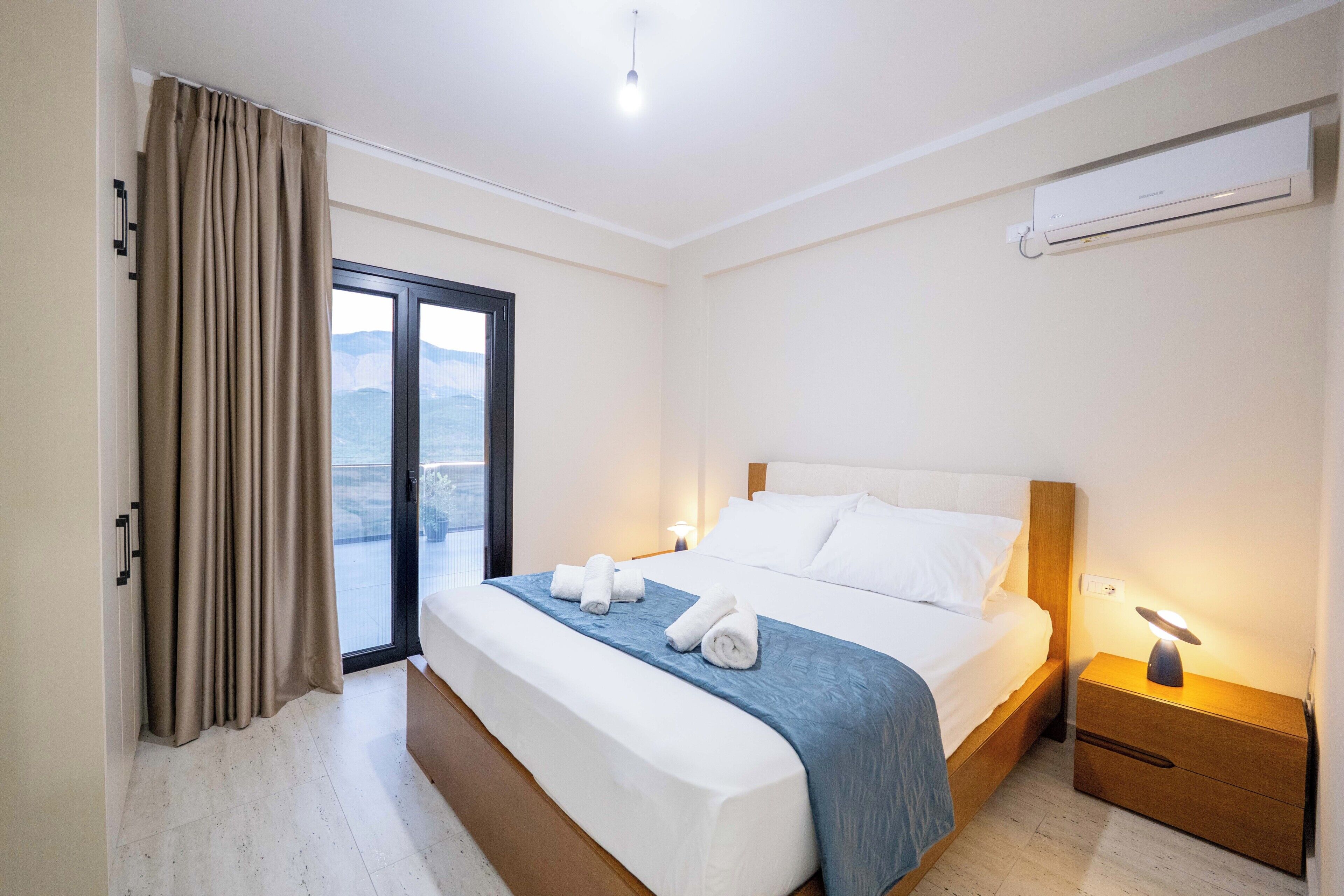 A hotel room with a bed, a nightstand, a window with curtains, and an air conditioning unit on the wall.