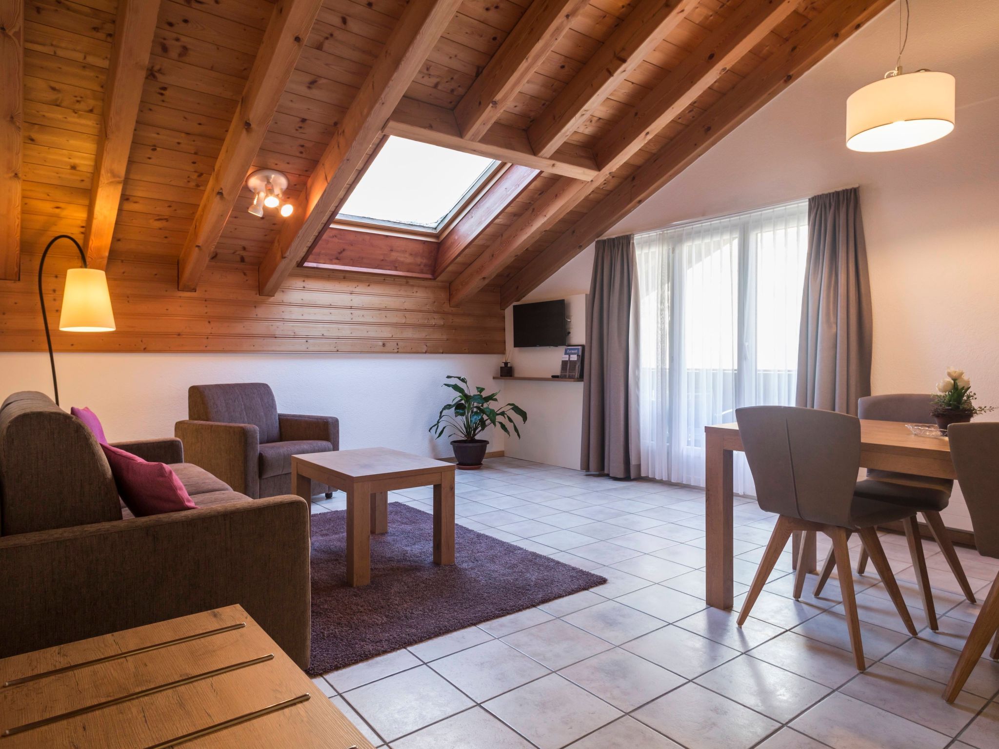 A cozy living room with a sloped ceiling, wooden beams, a flat-screen TV, a dining area with chairs, a coffee table, and a potted plant.