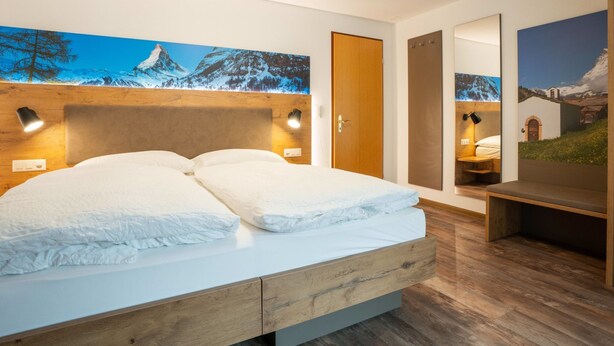Un dormitorio moderno con cabecera de madera, cama grande con ropa de cama blanca, dos lámparas de lectura fijadas a la pared y un espejo grande que refleja una vista panorámica.