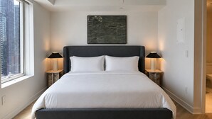 Quarto moderno com uma cama grande, duas luminárias de cabeceira, vista para a cidade e uma obra de arte emoldurada na parede.