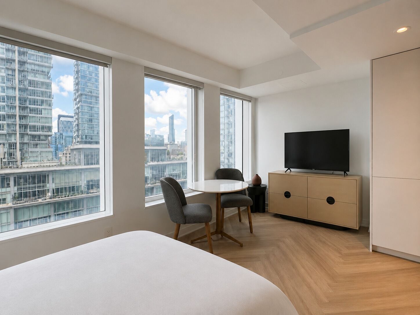 Een moderne hotelkamer met een groot bed, een eettafel, twee stoelen, een televisie op een kast en grote ramen met uitzicht over de stad.