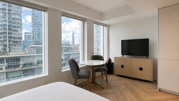 Een moderne hotelkamer met een groot bed, een eettafel, twee stoelen, een televisie op een kast en grote ramen met uitzicht over de stad.