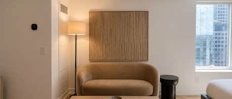 Habitación de hotel moderna con un sofá, una mesita de noche con libros, una lámpara y vista a la ciudad.