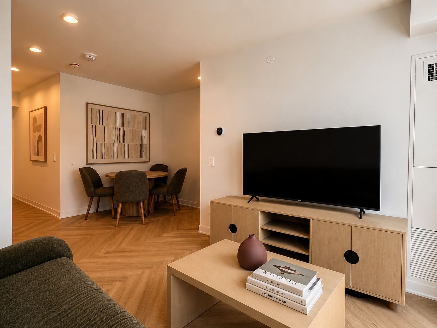 A modern living room with a flat-screen TV, a dining area with chairs, and a coffee table with books and a vase.