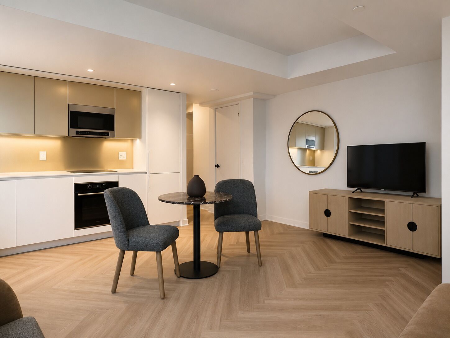 Una sala de estar moderna con un sofá, dos sillones, una mesa de centro, un televisor de pantalla plana y una zona de cocina con armarios y electrodomésticos.