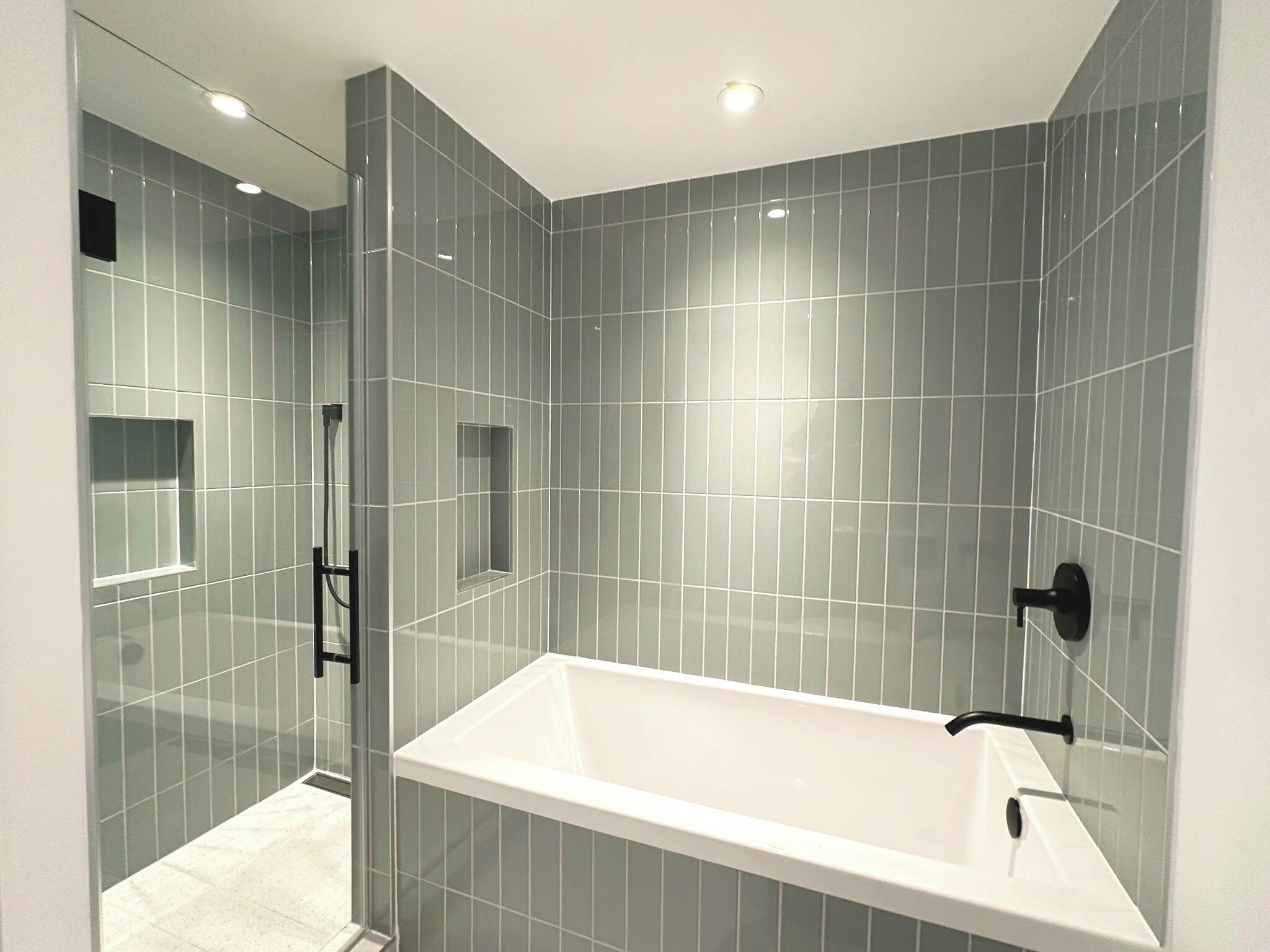 A modern bathroom with a glass shower enclosure, a white bathtub, and grey tiled walls.