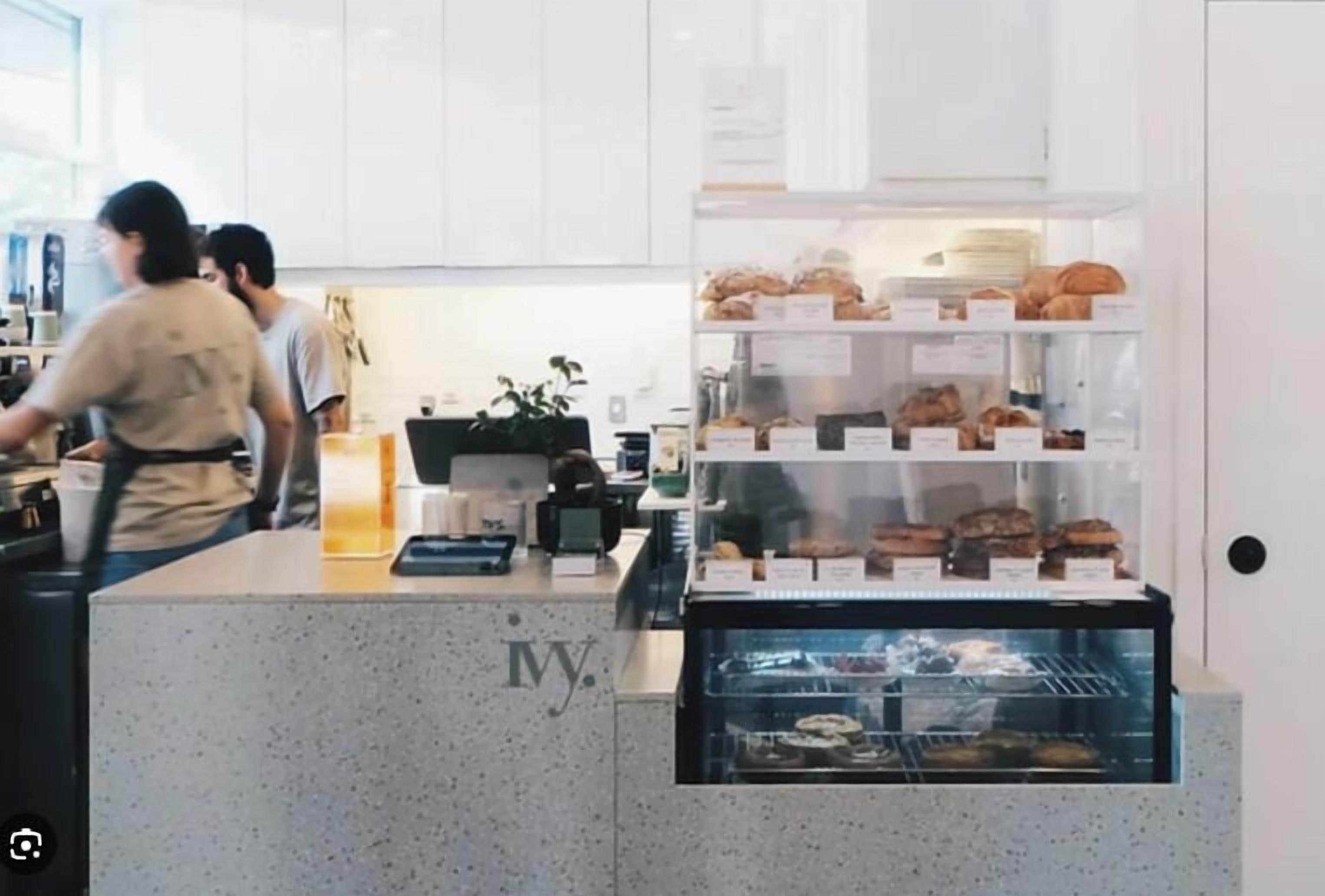 A bakery counter with pastries and a coffee machine, a person working behind the counter, and a display case with baked goods.