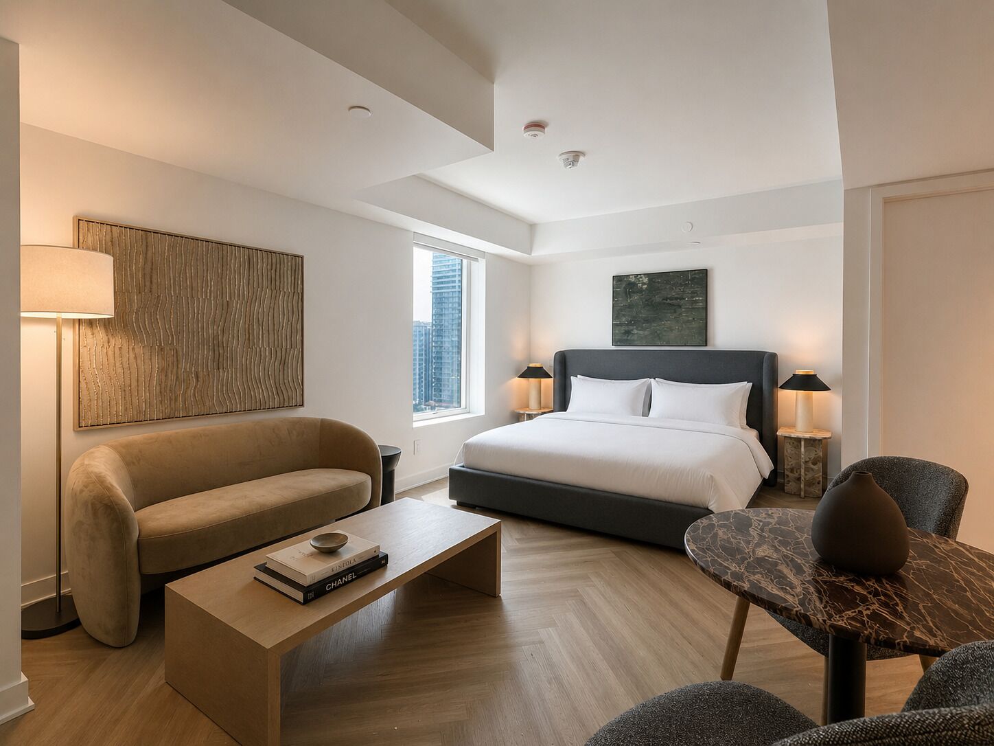 A modern hotel room with a large bed, a sofa, a coffee table with books, a side table, and a view of the city.