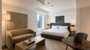 A modern hotel room with a large bed, a sofa, a coffee table with books, a side table, and a view of the city.