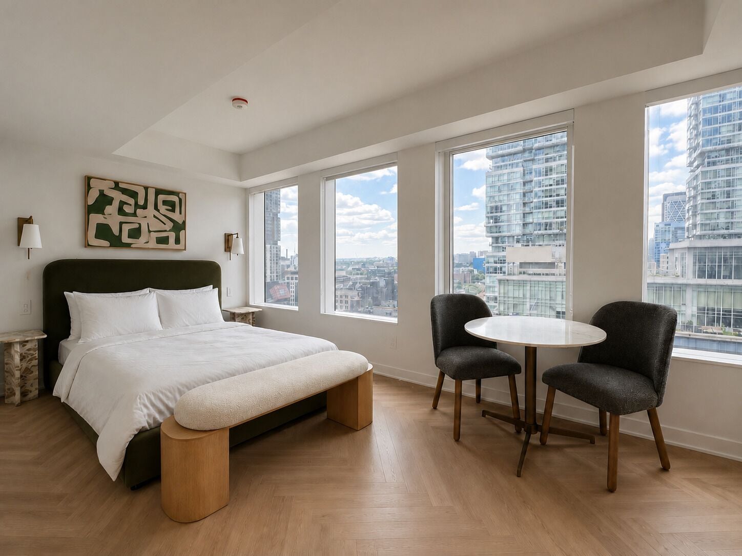 A modern hotel room with a large bed, two chairs, a small table, and a city view through large windows.