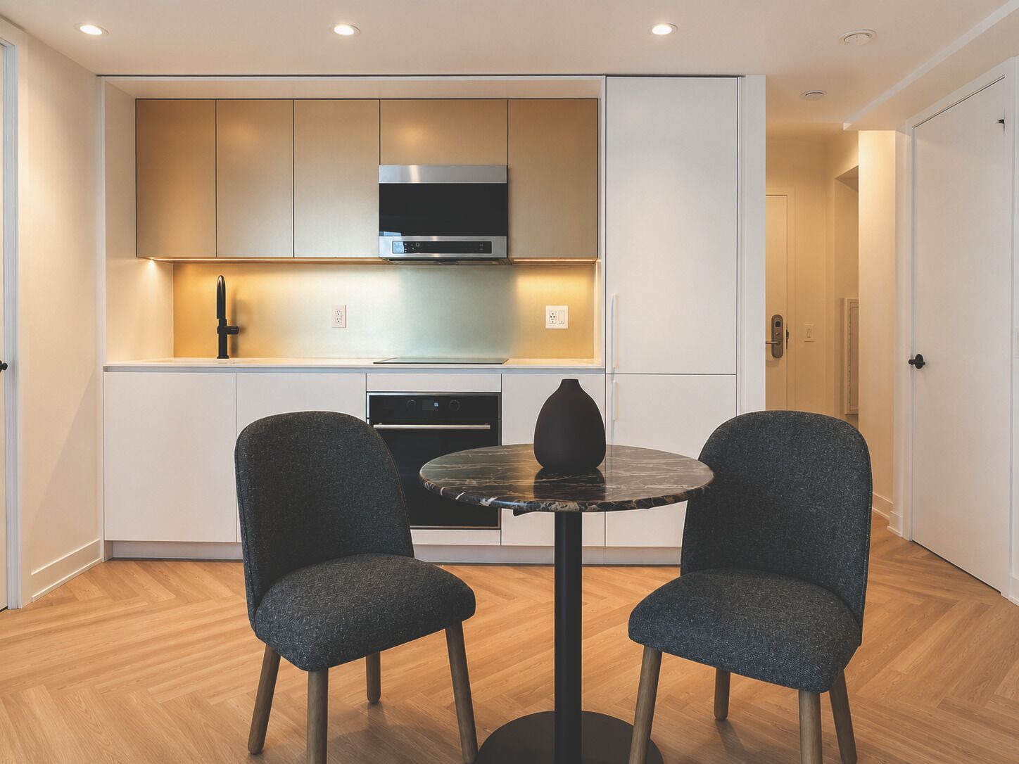 Una cocina moderna con horno empotrado y microondas, una mesa redonda con dos sillas, y una puerta que da a otra habitación.
