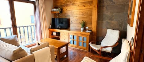 Una sala de estar acogedora con vigas de madera, un sofá, una mesa de centro y dos sillones. Un televisor está montado en la pared, y hay una chimenea de piedra.