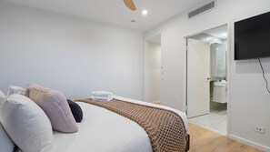Ein Schlafzimmer mit einem Bett, Kissen, einer Decke, einem Fernseher und einem durch eine offene Tür einsehbaren Badezimmer.