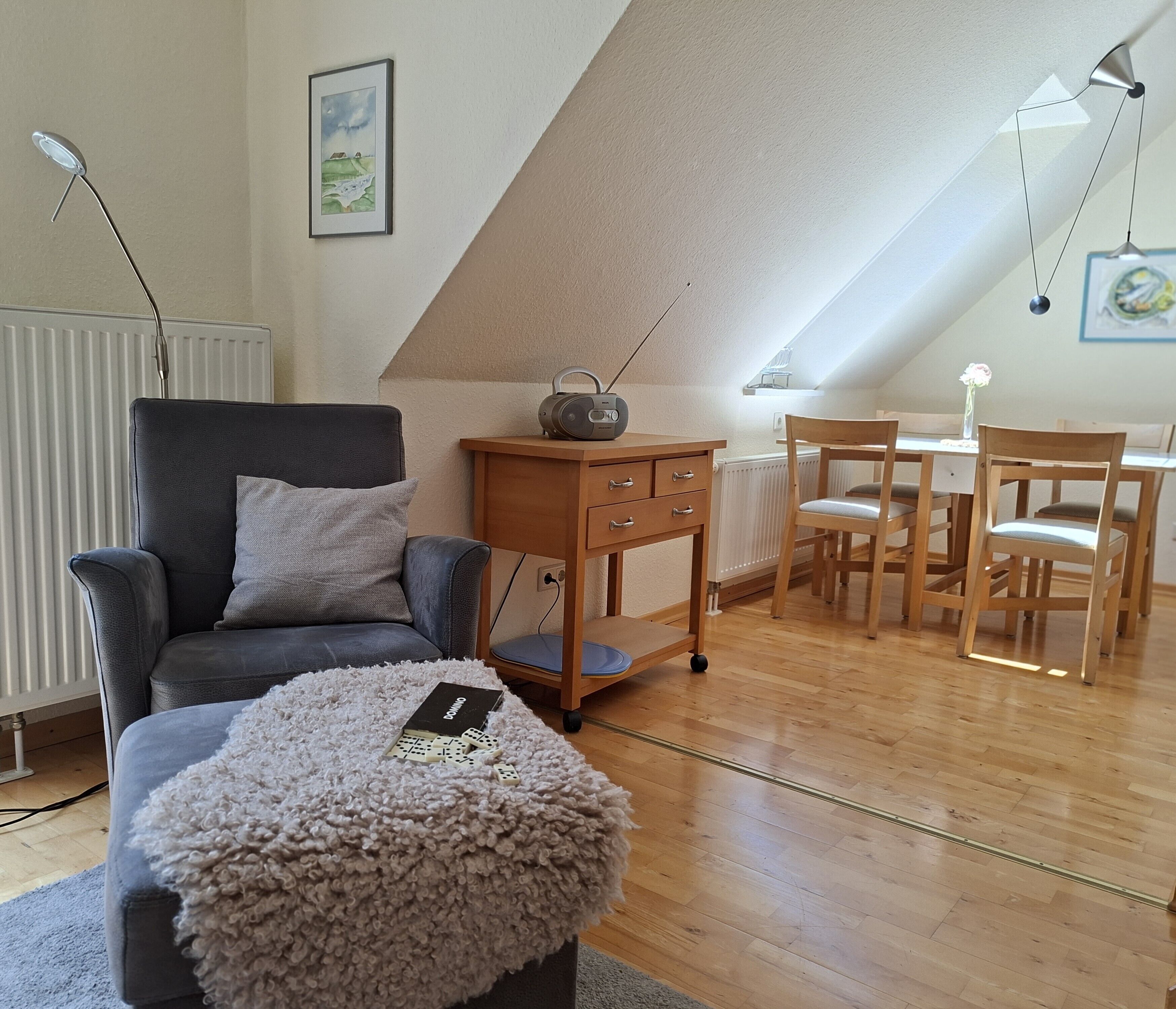 Una habitación acogedora con techo inclinado, un sillón gris, una mesita de madera con un radio, una mesa de comedor con sillas y un otoman suave.