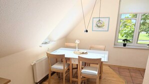 Uma área de refeições com mesa e cadeiras brancas, um radiador e uma janela com uma planta.