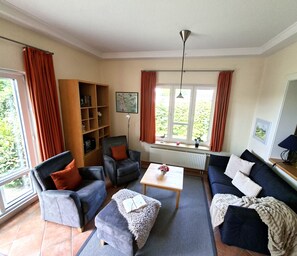 A living room with a sofa, chairs, a coffee table, a bookshelf, and a window with curtains. - High-quality "Strandläufer" vacation rental in Haus Wattenblick with its own sauna (Warwerort)