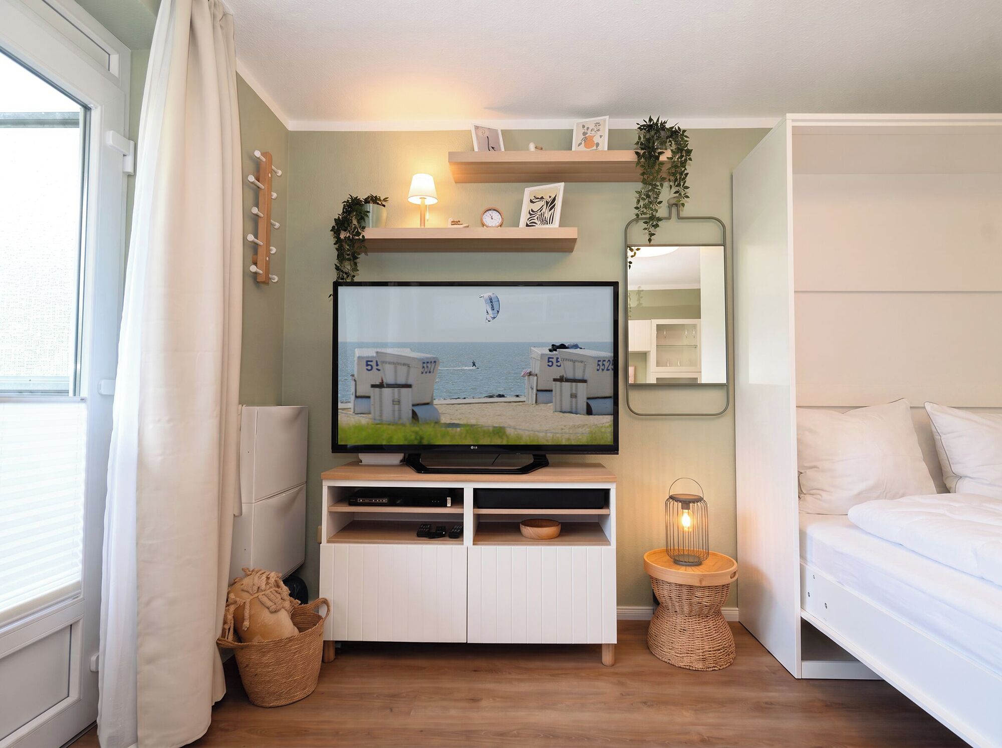 A modern bedroom with a television on a wooden stand, a bedside lamp, a mirror, and a window with curtains.