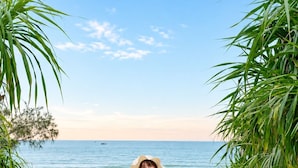 A beach scene with a person in a white dress holding a straw hat, walking on sandy terrain with vegetation on either side.