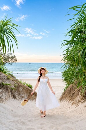 Escena de playa con una persona vestida de blanco sosteniendo un sombrero de paja, caminando sobre arena con vegetación a ambos lados.
