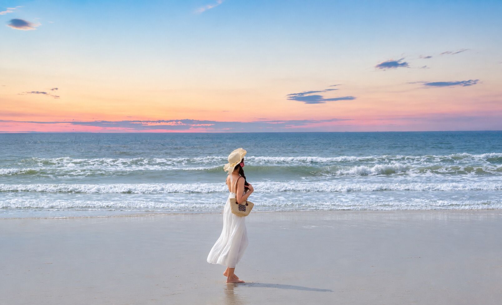 A woman in a white dress standing on a beach with the ocean and a sunset in the background.