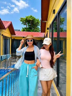Two individuals standing by a pool with a yellow building and red-tiled roof in the background.