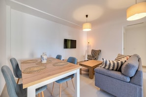 Ein modernes Wohnzimmer mit einer Couch, einem Esstisch mit Stühlen, einem an der Wand befestigten Fernseher und einer Hängelampe.