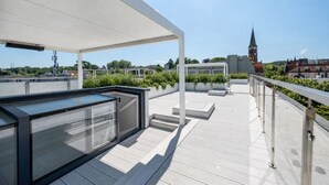 Une terrasse sur le toit, dotée d’une structure vitrée, d’espaces aménagés pour s’asseoir et offrant une vue sur le clocher d’une église au loin.