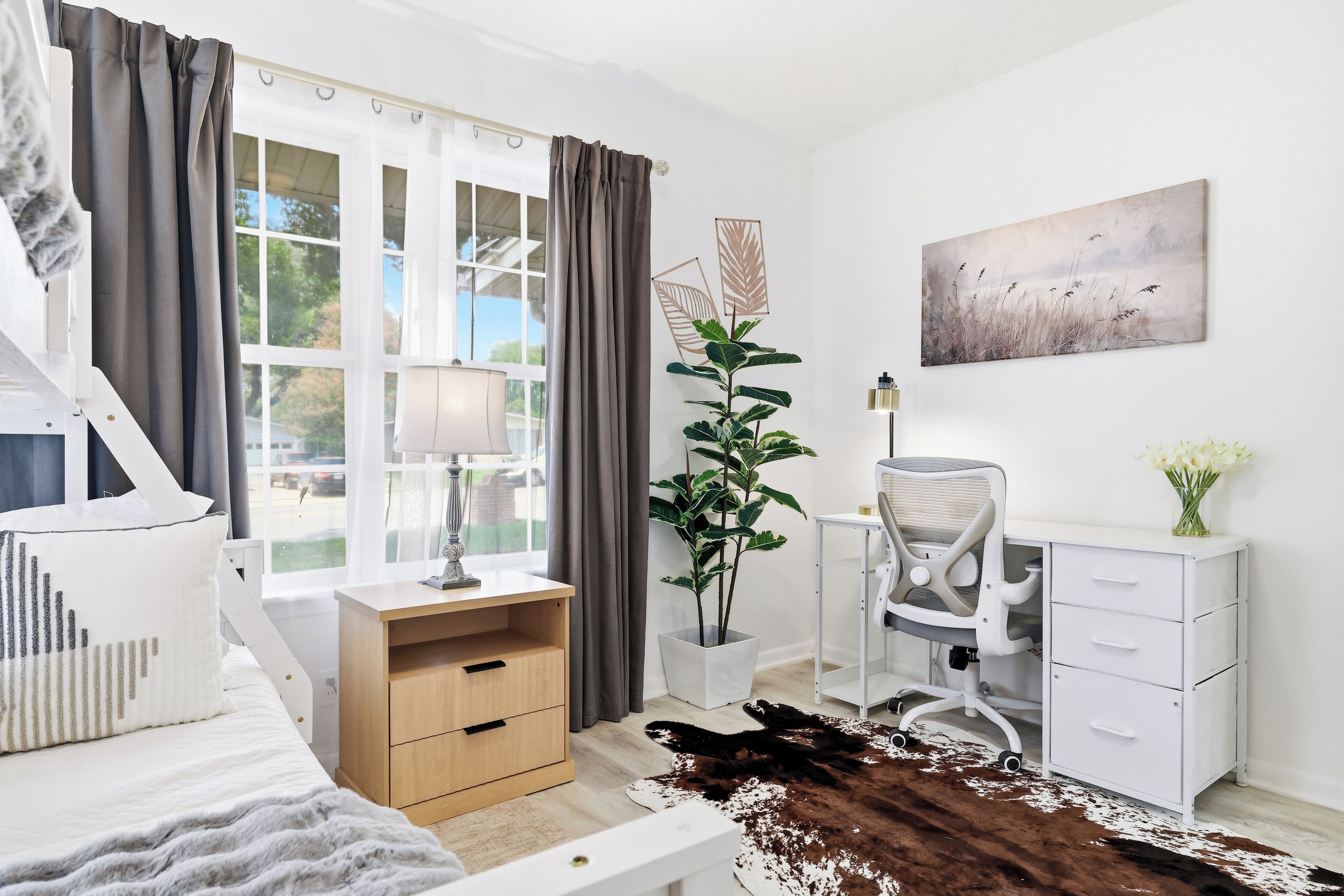 A modern bedroom with a bed, bedside table, lamp, desk, chair, potted plant, artwork, and a cowhide rug.