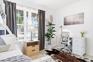 Un dormitorio moderno con cama, mesita de noche, lámpara, escritorio, silla, planta en maceta, obra de arte y alfombra de cuero de vaca.