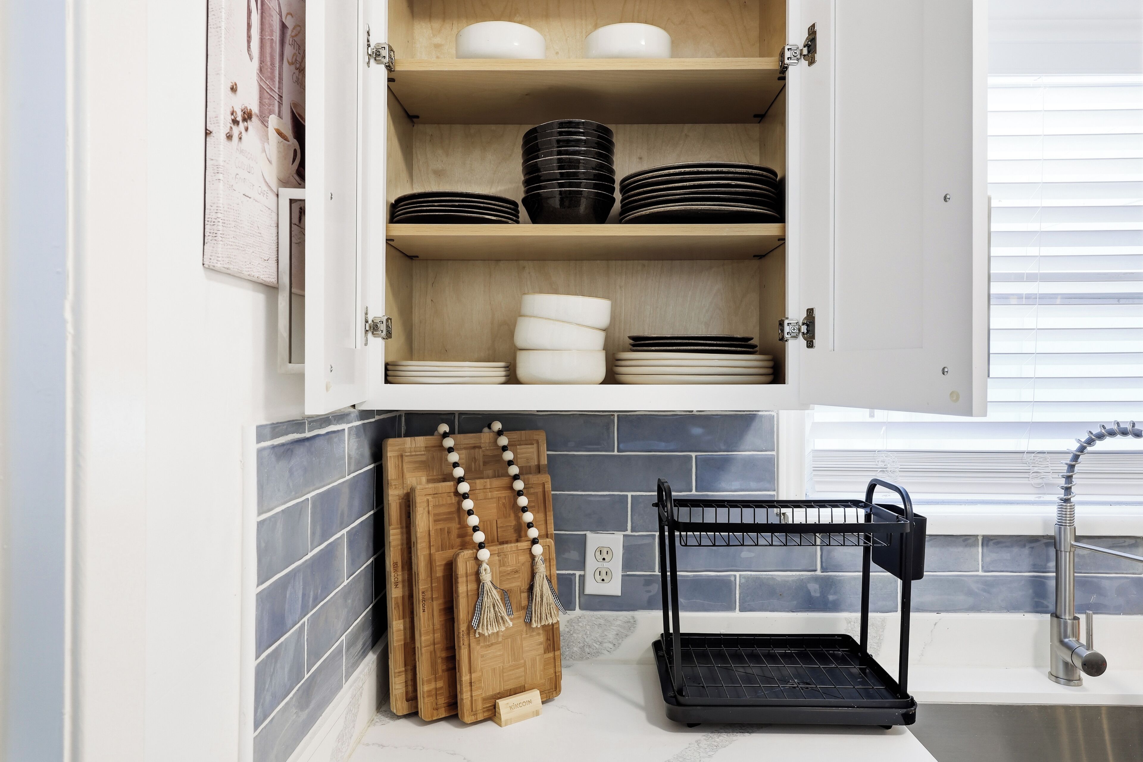 Una cocina con un armario que contiene platos y tazones, una tabla de cortar de madera y un escurreplatos negro.